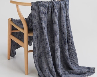 May include: A dark blue waffle-weave throw blanket draped over a light-colored wooden chair with a curved backrest and woven seat. The blanket features a distinct grid-like texture and hangs down to the floor in soft, vertical folds.