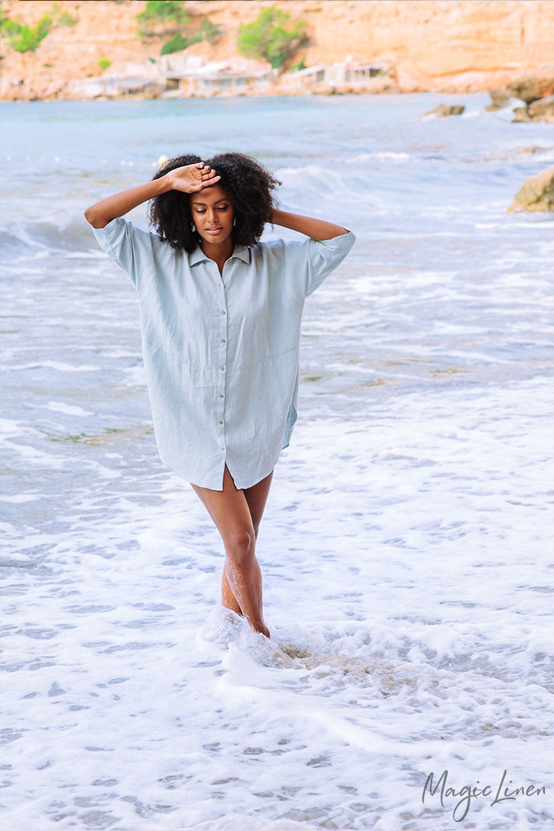 Pode incluir: Uma mulher vestindo um vestido camisa de linho azul claro est&aacute; de p&eacute; nas ondas do oceano. O vestido camisa tem gola e bot&otilde;es na frente. A mulher tem cabelo escuro e encaracolado e est&aacute; olhando para baixo.