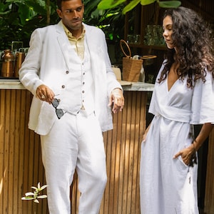May include: A man and a woman standing in front of a wooden bar. The man is wearing a white linen suit and the woman is wearing a white linen dress. They are both looking at each other.