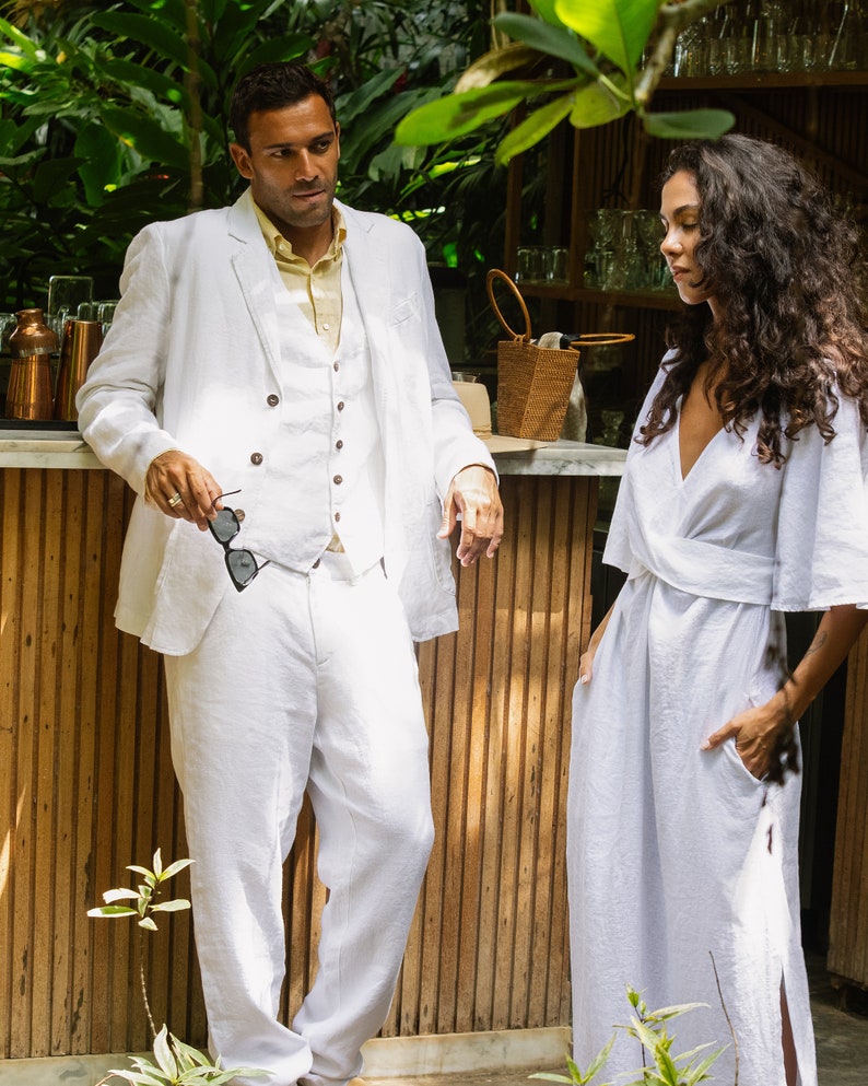 May include: A man and a woman standing in front of a wooden bar. The man is wearing a white linen suit and the woman is wearing a white linen dress. They are both looking at each other.
