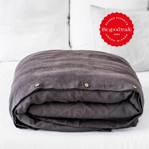 May include: A charcoal gray duvet cover featuring a row of three light-colored wooden buttons along the folded edge. A red circular badge with white text reads "READER FAVORITE the good trade 2024 AWARD WINNER" positioned in the upper right corner.