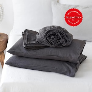 May include: A set of dark charcoal gray bed linens including two pillowcases and a fitted sheet, stacked on a white bed. A red circular badge in the upper right corner features the text: READER FAVORITE THE GOOD TRADE AWARD WINNER 2024.