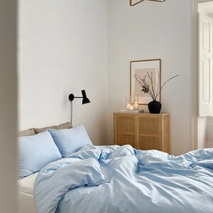 May include: A light blue duvet cover with white pillowcases on a bed in a bedroom with a white wall, a wooden side table with a wicker front, a framed print, and a black lamp on the wall.