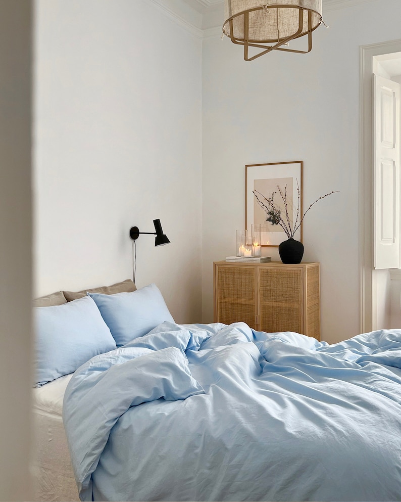 May include: A light blue duvet cover with white pillowcases on a bed in a bedroom with a white wall, a wooden side table with a wicker front, a framed print, and a black lamp on the wall.