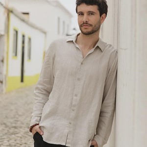 Pu&ograve; includere: Una camicia beige chiaro a maniche lunghe con bottoni. La camicia &egrave; realizzata in un tessuto leggero e ha un colletto classico. La persona indossa pantaloni neri e ha le mani in tasca. Lo sfondo &egrave; un muro bianco e una strada acciottolata.