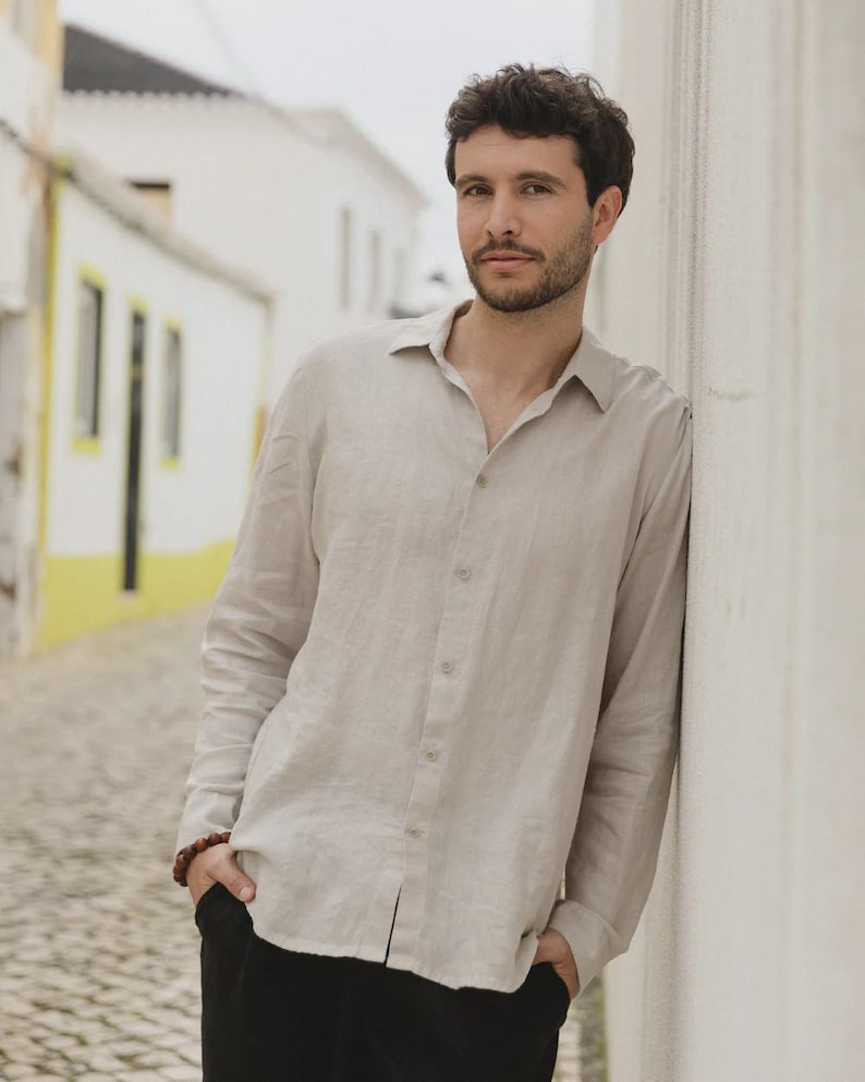 Pu&ograve; includere: Una camicia beige chiaro a maniche lunghe con bottoni. La camicia &egrave; realizzata in un tessuto leggero e ha un colletto classico. La persona indossa pantaloni neri e ha le mani in tasca. Lo sfondo &egrave; un muro bianco e una strada acciottolata.