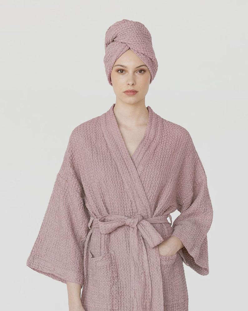 K&ouml;nnte beinhalten: Ein mauvefarbener Waffel-Bademantel und ein passendes Handtuch im Turban-Stil. Der Bademantel hat ein Wickeldesign mit Bindeg&uuml;rtel, Taschen und weiten &Auml;rmeln. Das Handtuch ist um den Kopf gewickelt. Der Stoff hat eine strukturierte Optik.