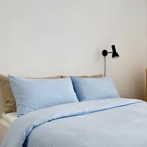 May include: A light blue bed with two pillows and a white wall in the background. A black wall sconce is mounted on the wall.