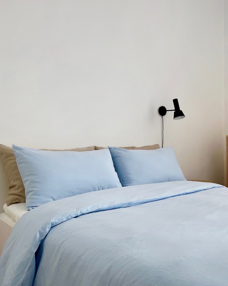 May include: A light blue bed with two pillows and a white wall in the background. A black wall sconce is mounted on the wall.