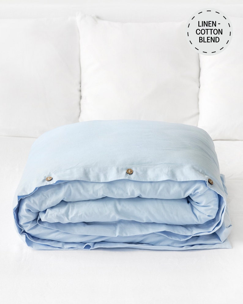 May include: A light blue linen-cotton blend duvet cover with three wooden buttons. The duvet cover is folded and placed on a white surface.