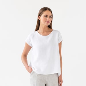 Può includere: Una blusa bianca in lino con maniche corte e scollo rotondo. La blusa è ampia e ha una forma leggermente a trapezio.