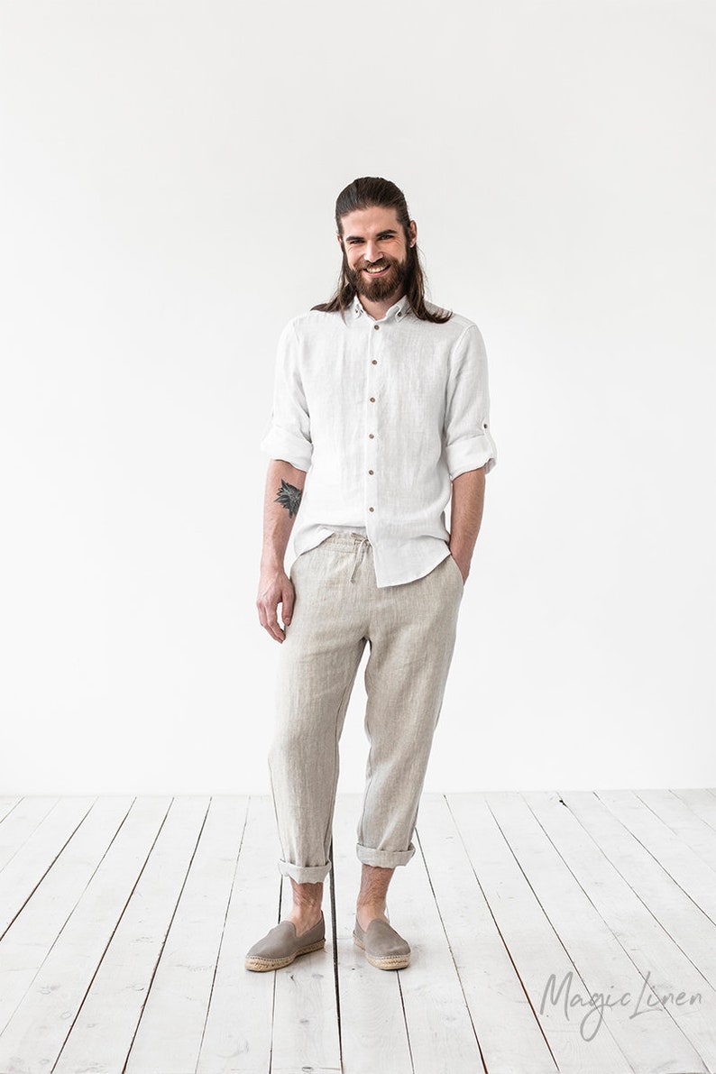 May include: A man wearing a white linen shirt and beige linen pants. He is standing on a wooden floor and smiling at the camera. The text "Magic Linen" is visible in the bottom right corner of the image.