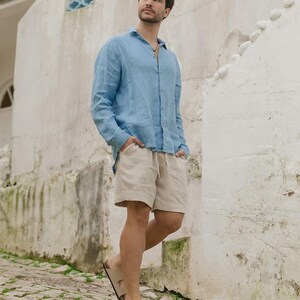 Pu&ograve; includere: Una camicia di lino azzurra e pantaloncini beige sono indossati con sandali beige. La camicia &egrave; abbottonata e i pantaloncini hanno un cordoncino. I sandali hanno due cinturini. La persona &egrave; in piedi su una strada acciottolata.
