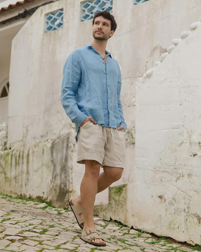 Pu&ograve; includere: Una camicia di lino azzurra e pantaloncini beige sono indossati con sandali beige. La camicia &egrave; abbottonata e i pantaloncini hanno un cordoncino. I sandali hanno due cinturini. La persona &egrave; in piedi su una strada acciottolata.