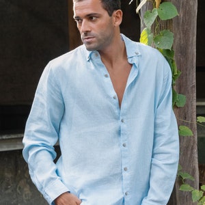 Pu&ograve; includere: Una camicia di lino blu chiaro con colletto abbottonato e maniche lunghe. La camicia &egrave; sbottonata in alto, rivelando il petto della persona che la indossa.