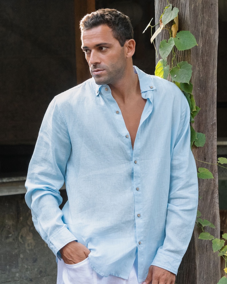 Pu&ograve; includere: Una camicia di lino blu chiaro con colletto abbottonato e maniche lunghe. La camicia &egrave; sbottonata in alto, rivelando il petto della persona che la indossa.