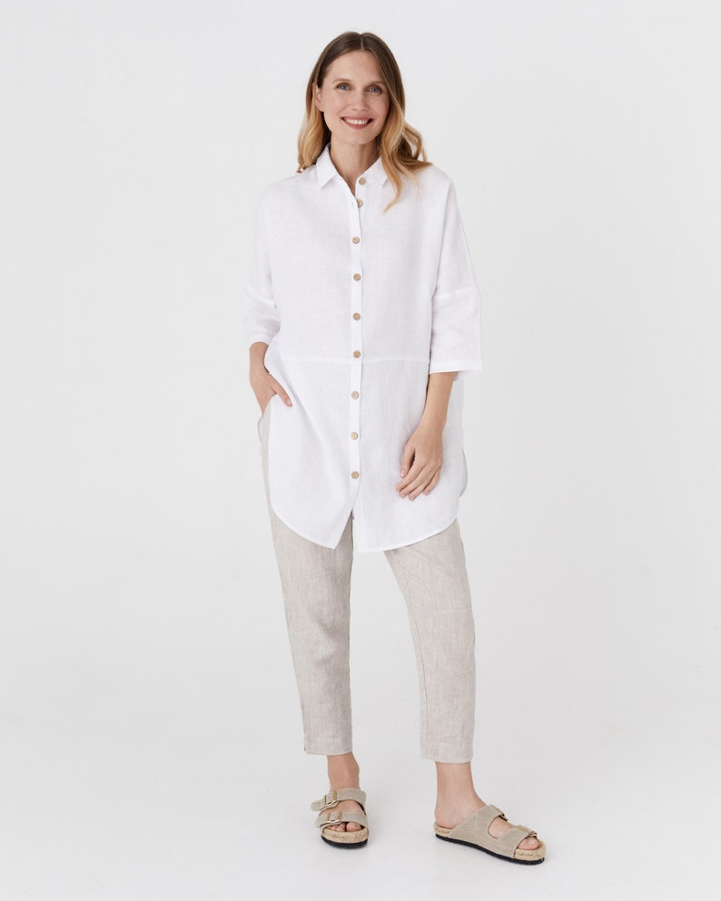 Pode incluir: Uma camisa branca de linho com colarinho de bot&otilde;es e mangas tr&ecirc;s quartos. A camisa &eacute; larga e tem um comprimento mais longo. A modelo est&aacute; usando a camisa com cal&ccedil;as de linho bege e sand&aacute;lias marrons.