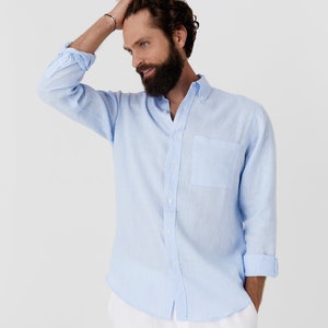 K&ouml;nnte beinhalten: Hellblaues Button-Down-Hemd mit Brusttasche und hochgekrempelten &Auml;rmeln. Das Hemd ist aus leichtem Stoff. Das Model tr&auml;gt eine wei&szlig;e Hose.