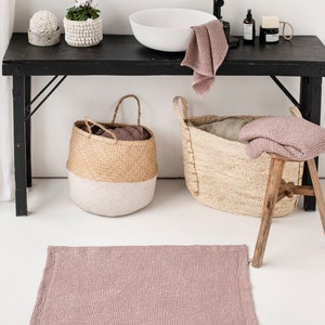 May include: A pink waffle weave bath mat in a bathroom setting. The mat is on a white floor in front of a black table with a white sink, two baskets, and plants.