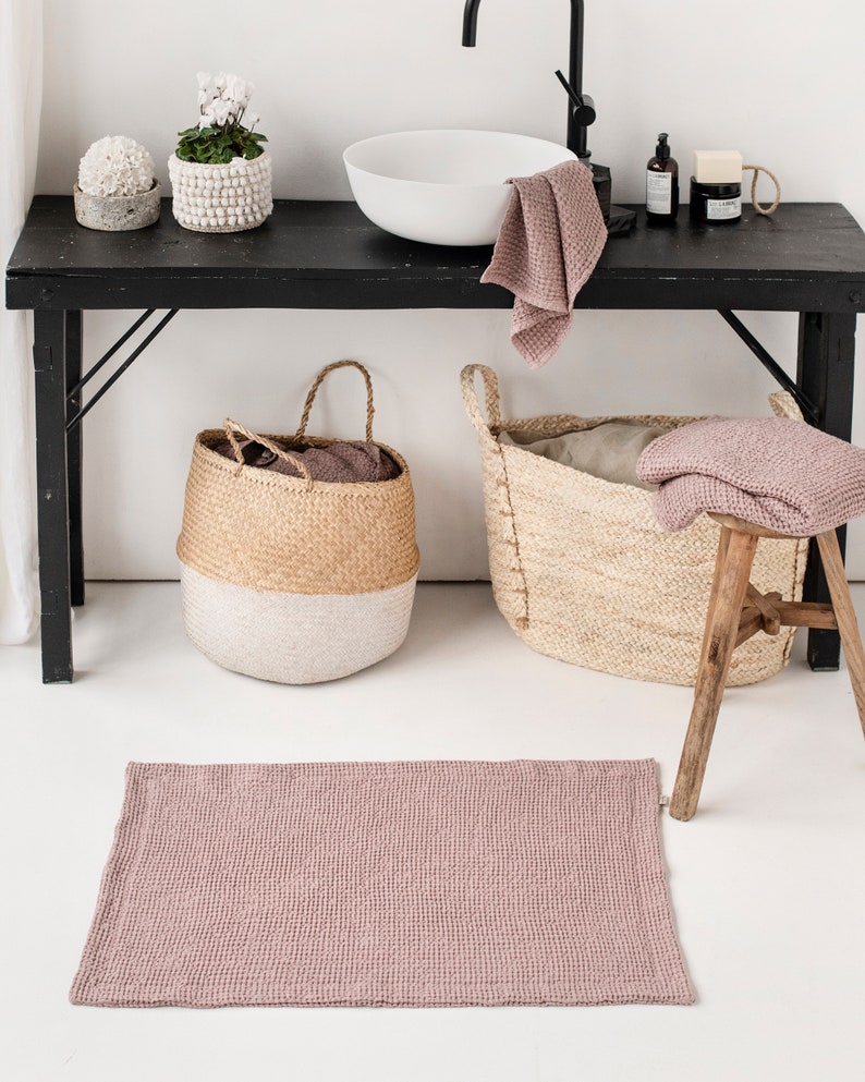 May include: A pink waffle weave bath mat in a bathroom setting. The mat is on a white floor in front of a black table with a white sink, two baskets, and plants.
