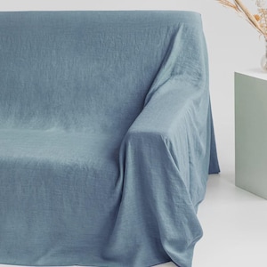 Könnte beinhalten: Eine hellblaue Sofahusse, die über ein Sofa drapiert ist. Die Hülle hat ein strukturiertes Aussehen und besteht aus einem weichen Stoff. Das Sofa steht vor einem weißen Hintergrund, mit einem kleinen Beistelltisch und getrockneten Blumen.