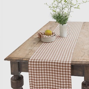 Puede incluir: Un camino de mesa a cuadros marrón y blanco sobre una mesa de madera. Una jarra blanca con ramitas verdes y pequeñas flores blancas está sobre la mesa. Un cuenco pequeño con una servilleta doblada y una pera amarilla también está sobre la mesa.