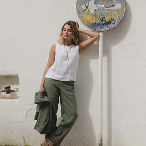 May include: A woman wearing a white sleeveless top and olive green pants. She is holding a matching jacket. The outfit is paired with beige wedge sandals. A necklace with a pendant is visible. The background features a white wall and a street sign.