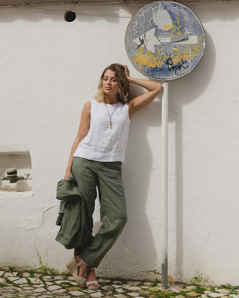 May include: A woman wearing a white sleeveless top and olive green pants. She is holding a matching jacket. The outfit is paired with beige wedge sandals. A necklace with a pendant is visible. The background features a white wall and a street sign.