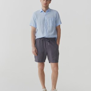 May include: A person wearing a light blue short-sleeve button-down shirt and charcoal gray shorts with a drawstring waist. The person is also wearing off-white sneakers.