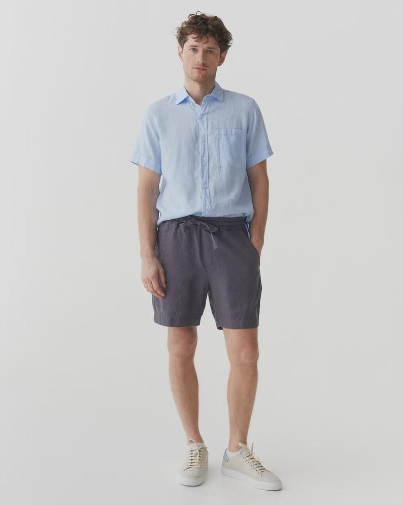 May include: A person wearing a light blue short-sleeve button-down shirt and charcoal gray shorts with a drawstring waist. The person is also wearing off-white sneakers.