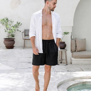 May include: A person wearing a white linen shirt and black shorts. The shirt is unbuttoned, and the shorts have a drawstring. The person is walking barefoot on a light-colored surface near a pool.