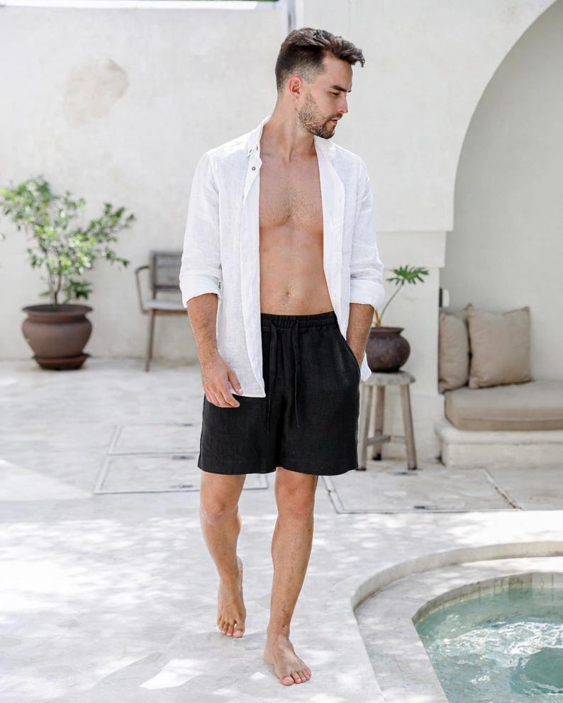 May include: A person wearing a white linen shirt and black shorts. The shirt is unbuttoned, and the shorts have a drawstring. The person is walking barefoot on a light-colored surface near a pool.