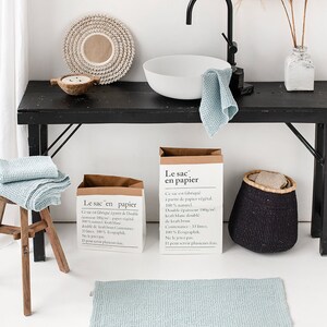May include: Two brown paper bags with the text "Le sac en papier" printed on them. The bags are sitting on a black table with a white ceramic sink, a blue towel, and a black woven basket. The text on the bags reads: "Ce sac est fabriqu&eacute; &agrave; partir de Double &eacute;paisseur 180g/m&sup2; papier v&eacute;g&eacute;tal. 100% naturel. Kraft blanc doubl&eacute; de kraft brun. Contenance: 33 litres. 100% Ecographik. Ne le jetez pas. Il peut servir plusieurs fois."