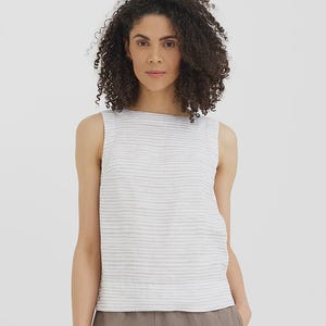 May include: Sleeveless white and gray striped top. The top has a round neckline and a textured appearance. The model is wearing the top with brown pants. The top is made of a lightweight fabric.