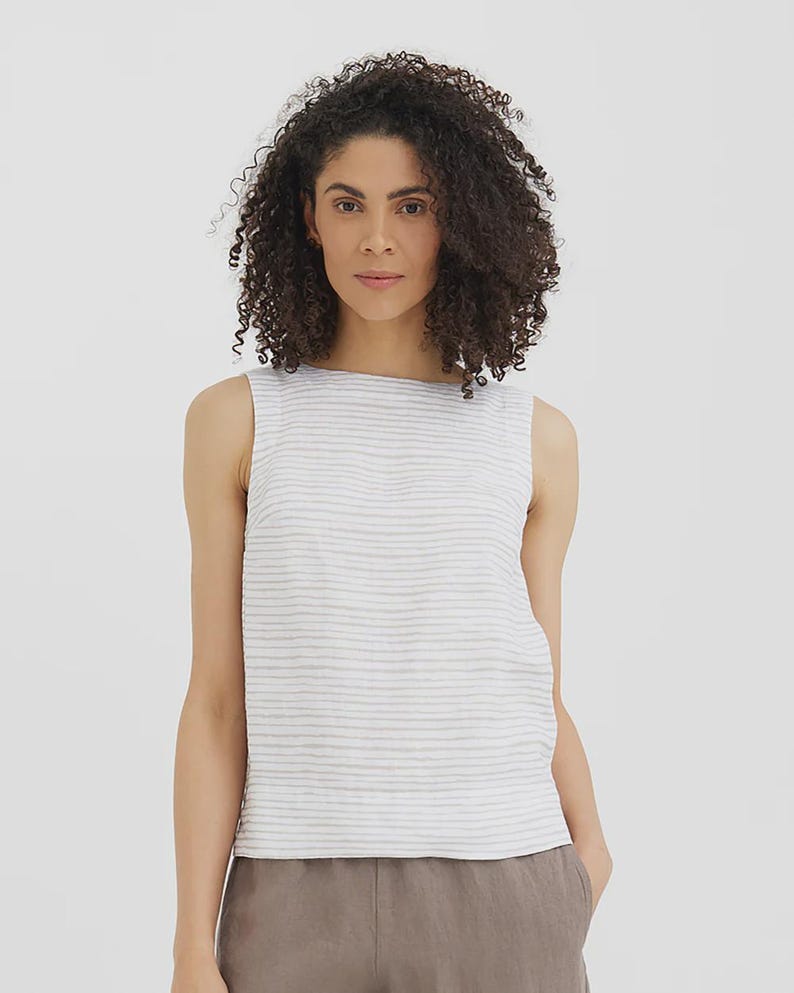 May include: Sleeveless white and gray striped top. The top has a round neckline and a textured appearance. The model is wearing the top with brown pants. The top is made of a lightweight fabric.