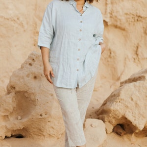 Pode incluir: Uma mulher vestindo uma camisa de linho azul claro com bot&otilde;es e cal&ccedil;as de linho combinando. Ela est&aacute; em p&eacute; em uma &aacute;rea arenosa com grandes rochas ao fundo.