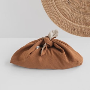 May include: A brown linen fabric wrap tied in a knot, with a light beige interior visible at the knot. The wrap is on a white surface, with a woven circular wall hanging in the background.