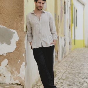 Pu&ograve; includere: Una camicia beige chiaro di lino e pantaloni neri. La camicia a maniche lunghe &egrave; abbottonata in alto. I pantaloni sono ampi. La persona indossa sandali. Lo sfondo &egrave; un muro strutturato.