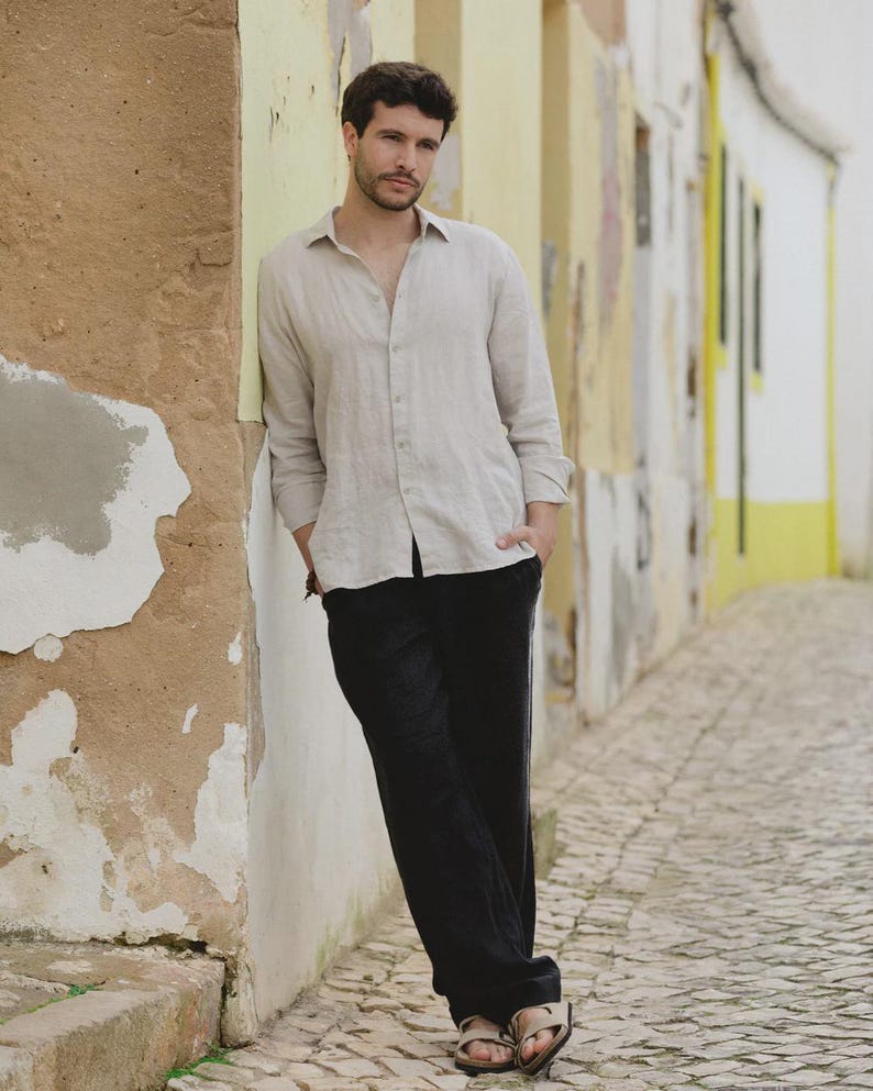 Pu&ograve; includere: Una camicia beige chiaro di lino e pantaloni neri. La camicia a maniche lunghe &egrave; abbottonata in alto. I pantaloni sono ampi. La persona indossa sandali. Lo sfondo &egrave; un muro strutturato.