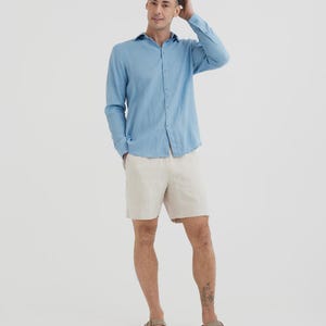 Pu&ograve; includere: Una camicia azzurra a maniche lunghe con bottoni e pantaloncini beige. La camicia &egrave; realizzata in tessuto leggero. I pantaloncini sono al ginocchio. La persona indossa sandali marroni.