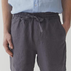 May include: Charcoal gray linen shorts with a drawstring waist. The shorts have a relaxed fit and pockets. The model is wearing a light blue button-down shirt. The shorts are made of a lightweight fabric.