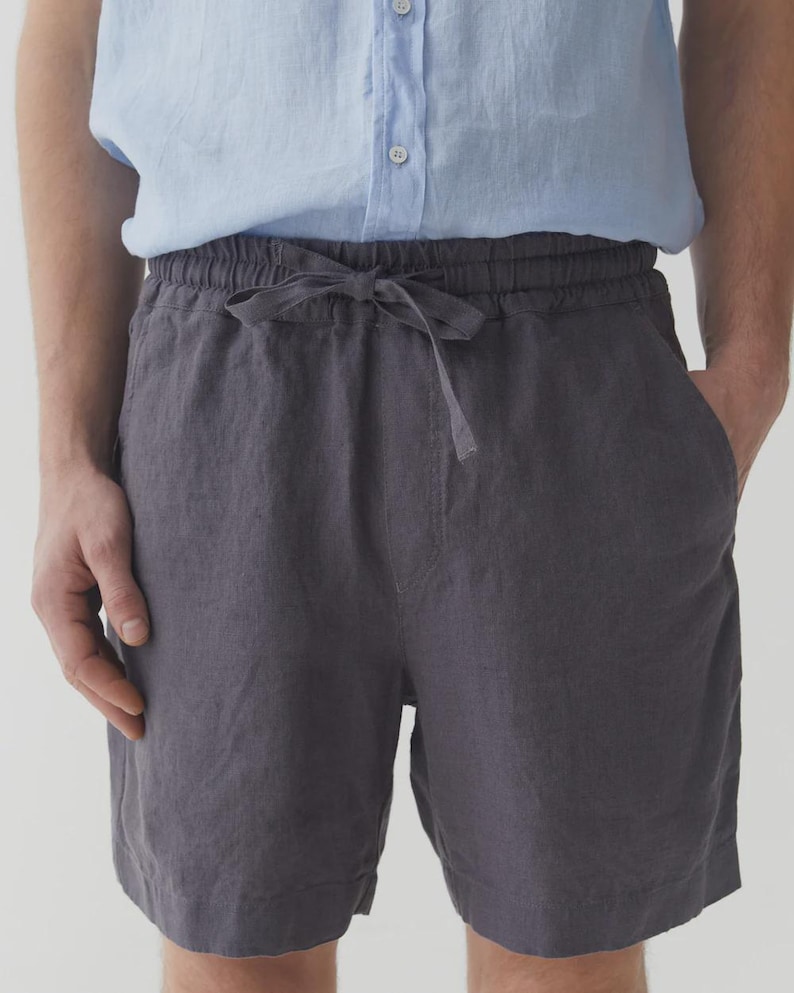 May include: Charcoal gray linen shorts with a drawstring waist. The shorts have a relaxed fit and pockets. The model is wearing a light blue button-down shirt. The shorts are made of a lightweight fabric.
