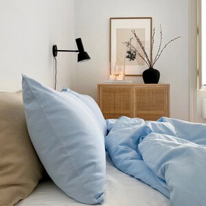 May include: A light blue bed with two pillows, one beige and one light blue, with a white bedspread. A wooden nightstand with two drawers is to the right of the bed, with a black vase with branches and candles on top. A wall-mounted black lamp is on the wall behind the bed.