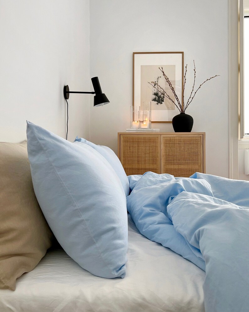May include: A light blue bed with two pillows, one beige and one light blue, with a white bedspread. A wooden nightstand with two drawers is to the right of the bed, with a black vase with branches and candles on top. A wall-mounted black lamp is on the wall behind the bed.