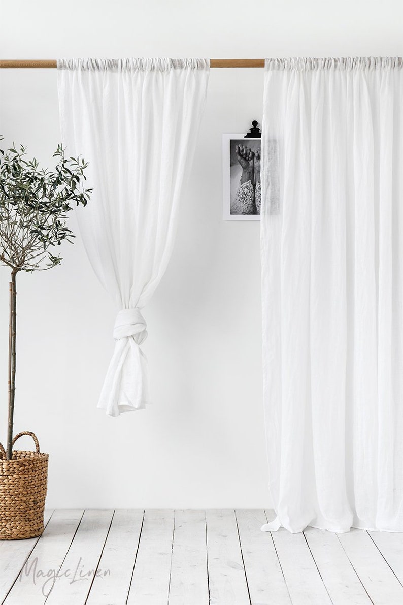 May include: Two white sheer linen curtains hanging from a wooden rod. The curtains are gathered at the top and tied back on one side. The curtains are in front of a white wall with a black and white picture hanging on it. A small potted olive tree is in the foreground. The floor is white wood.