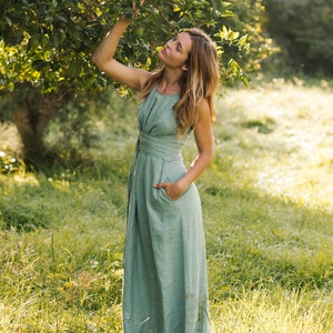 May include: A floor-length sage green sleeveless dress featuring a high neckline, a fitted bodice with a wide waistband, and a full-length skirt with side pockets. The garment is constructed from a solid-colored fabric with a visible natural texture.