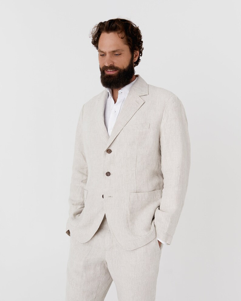 May include: A light beige linen suit jacket with a white button-down shirt underneath. The jacket has a single-breasted closure with four buttons and two flap pockets.
