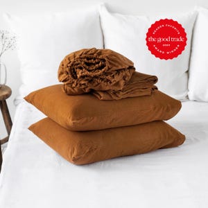 Peut inclure: Un ensemble de deux taies d'oreiller carrées de couleur rouille empilées sur un lit blanc, surmontées d'un drap-housse plié de couleur rouille. Un badge circulaire rouge dans le coin supérieur droit porte le texte : READER FAVORITE the good trade 2024 AWARD WINNER.