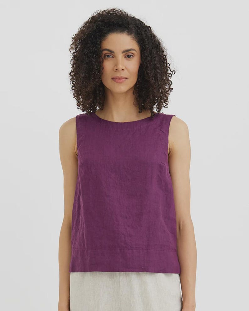 May include: A sleeveless, plum-colored top with a round neckline. The top is made of a textured fabric and has a simple, minimalist design. The model is wearing the top with light-colored pants.