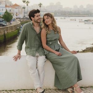 May include: An olive green long-sleeved button-down shirt, light beige trousers, and an olive green sleeveless maxi dress with a tie-waist detail. The clothing is worn with beige sandals and a wedge-heeled sandal, showcasing a coordinated set of apparel.